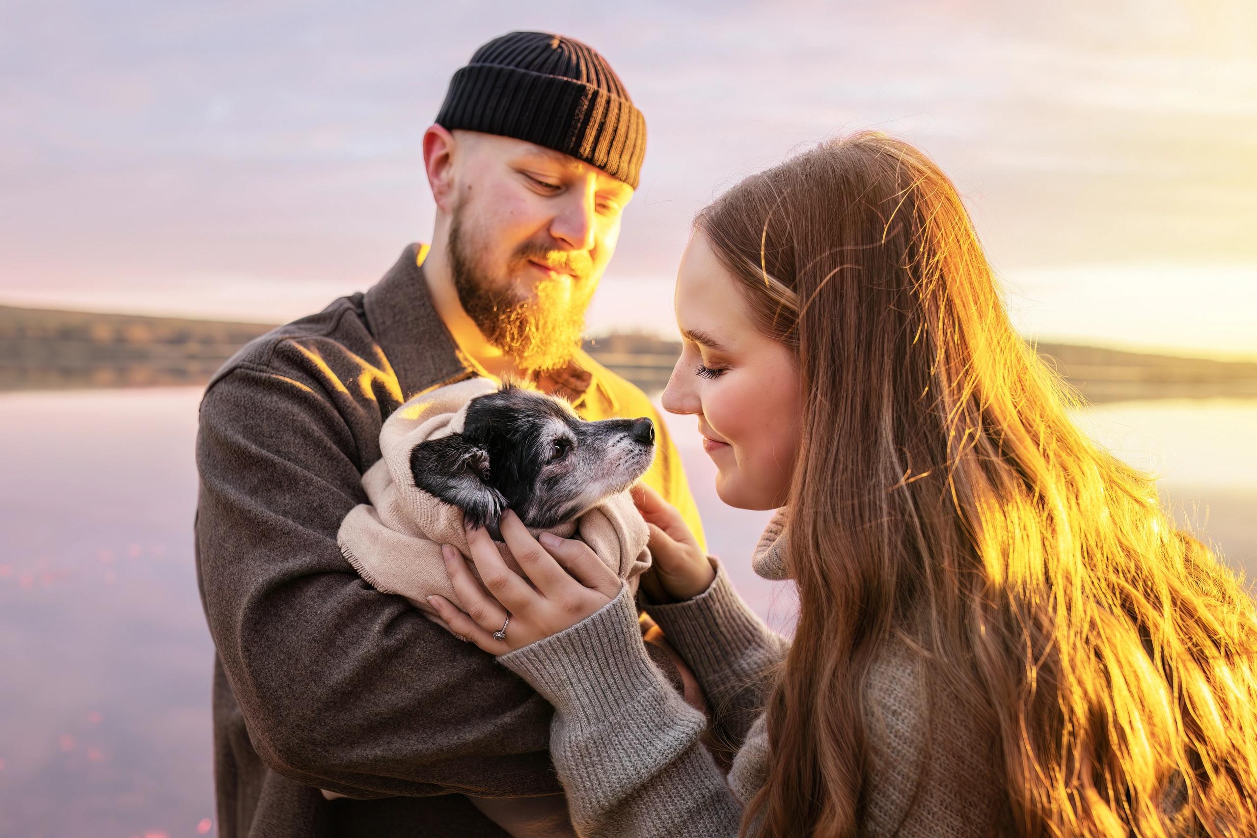 A couple holding their small dog in their arms by a lake at sunset